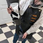 An orthotics practitioner in a denim work apron and powered full-face respirator holds a disassembled carbon fiber KAFO, showing the thigh section, knee joint, and foot plate.