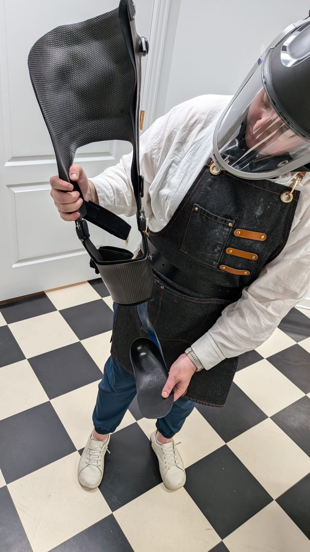 An orthotics practitioner in a denim work apron and powered full-face respirator holds a disassembled carbon fiber KAFO, showing the thigh section, knee joint, and foot plate.