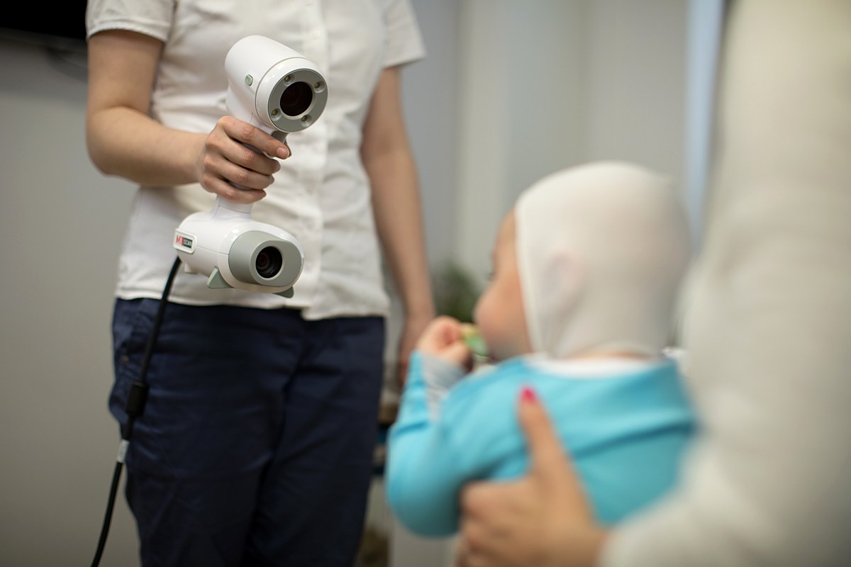 Baby wearing a scanning cap while a clinician holds a handheld 3D scanner for a flat head scan